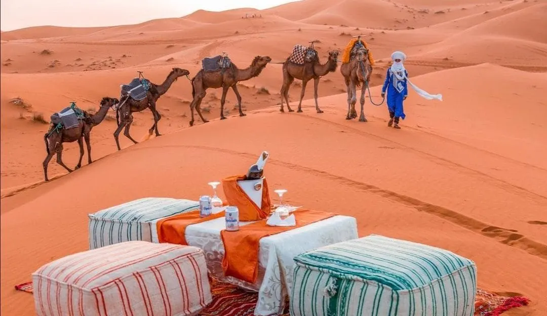 Cushioned seating at a desert camp with a camel caravan passing behind