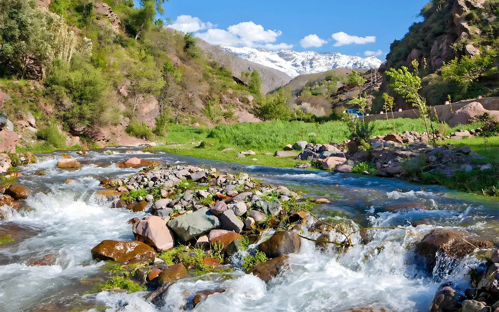 A rocky river in a green valley with snow-covered Atlas peaks behind